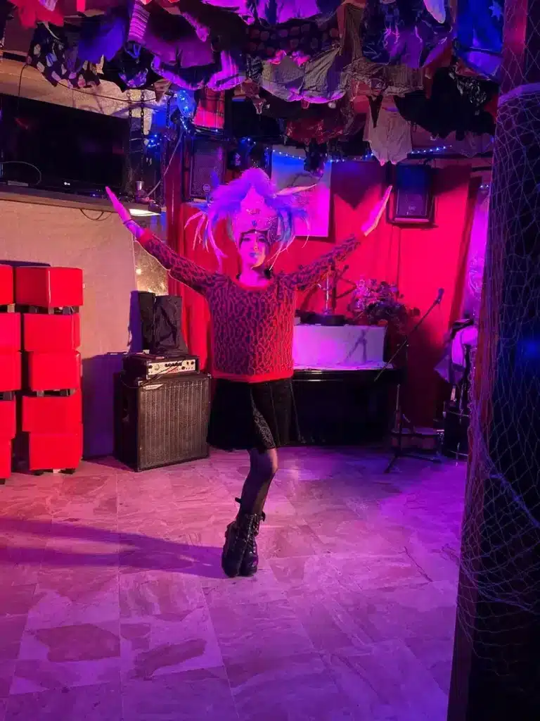 Performeuse en tenue léopard: ambiance magenta et décor de club Performeuse en tenue léopard et coiffe à plumes sur scène, bras levés. L'éclairage magenta illumine le club décoré de sous-vêtements suspendus.